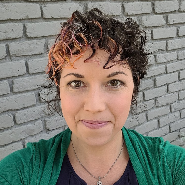 Woman with curly hair, head and shoulders, in front of grey brick wall